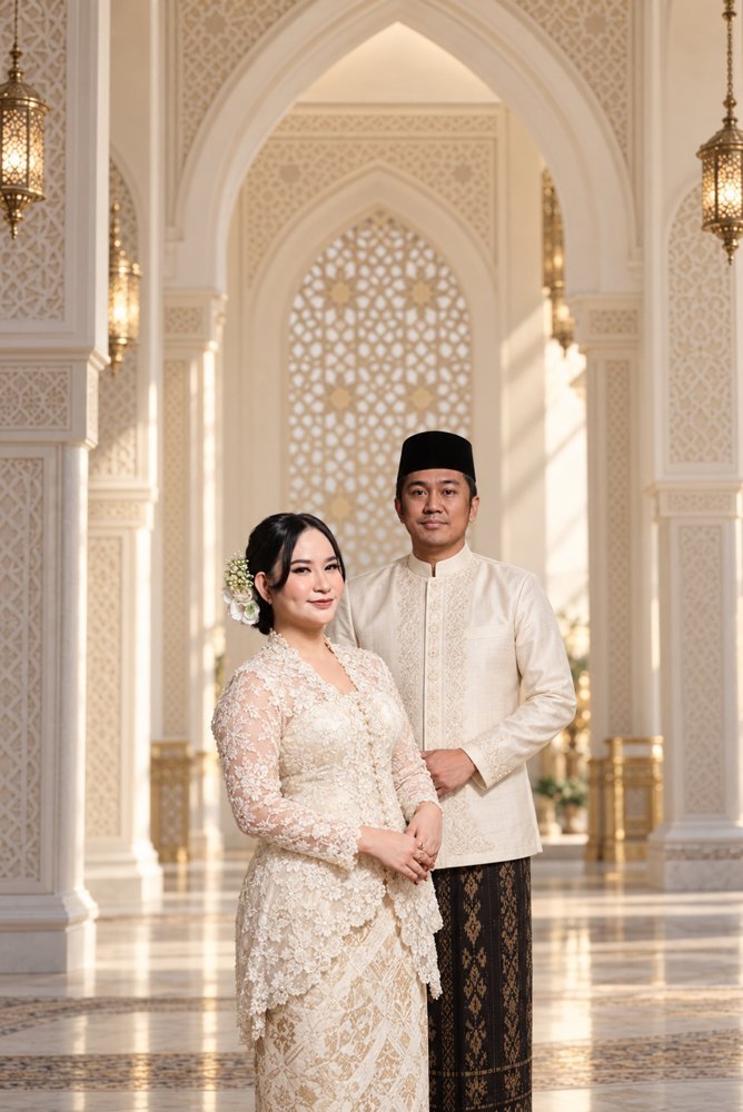 Iwan dan Yuli berdiri dalam busana putih dengan latar lengkungan emas